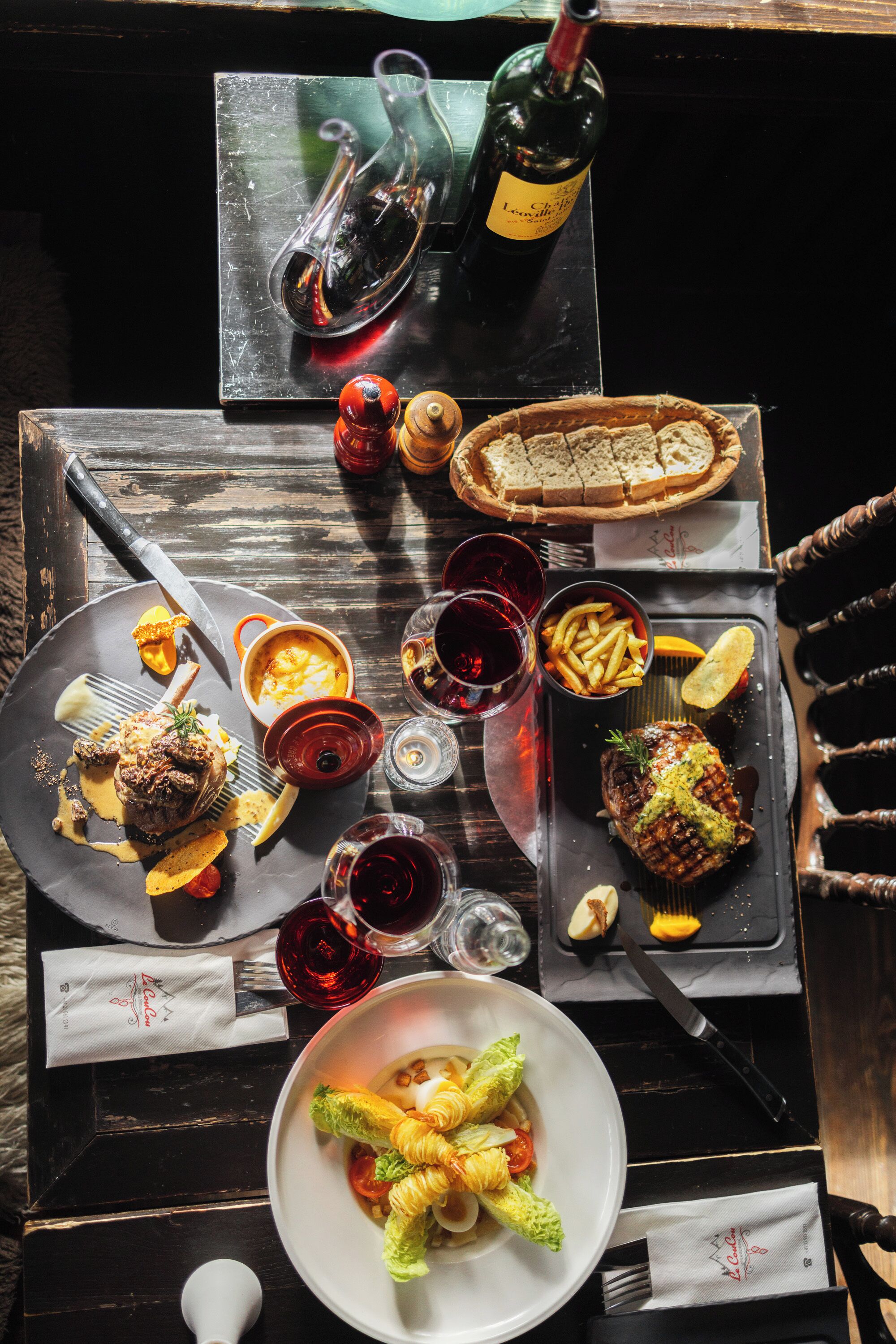 Een eettafel met diverse gerechten, waaronder steak, friet en salade, plus wijnkaraffen en een fles wijn.