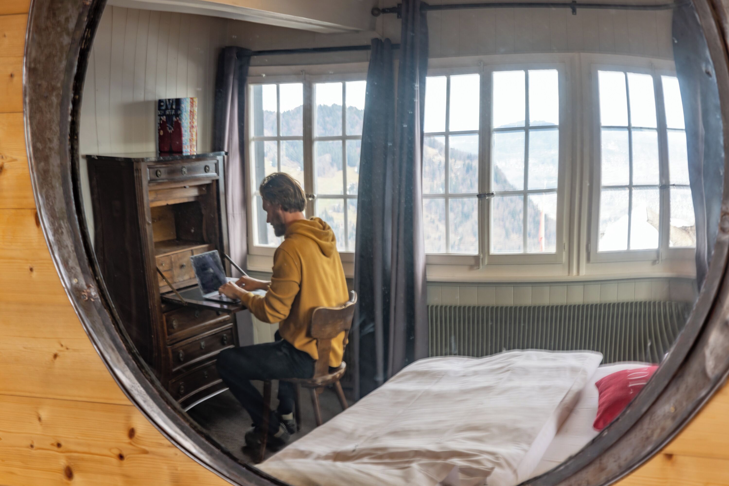 A room with a bed, a desk with a laptop, a window with curtains, and a view of the outdoors.