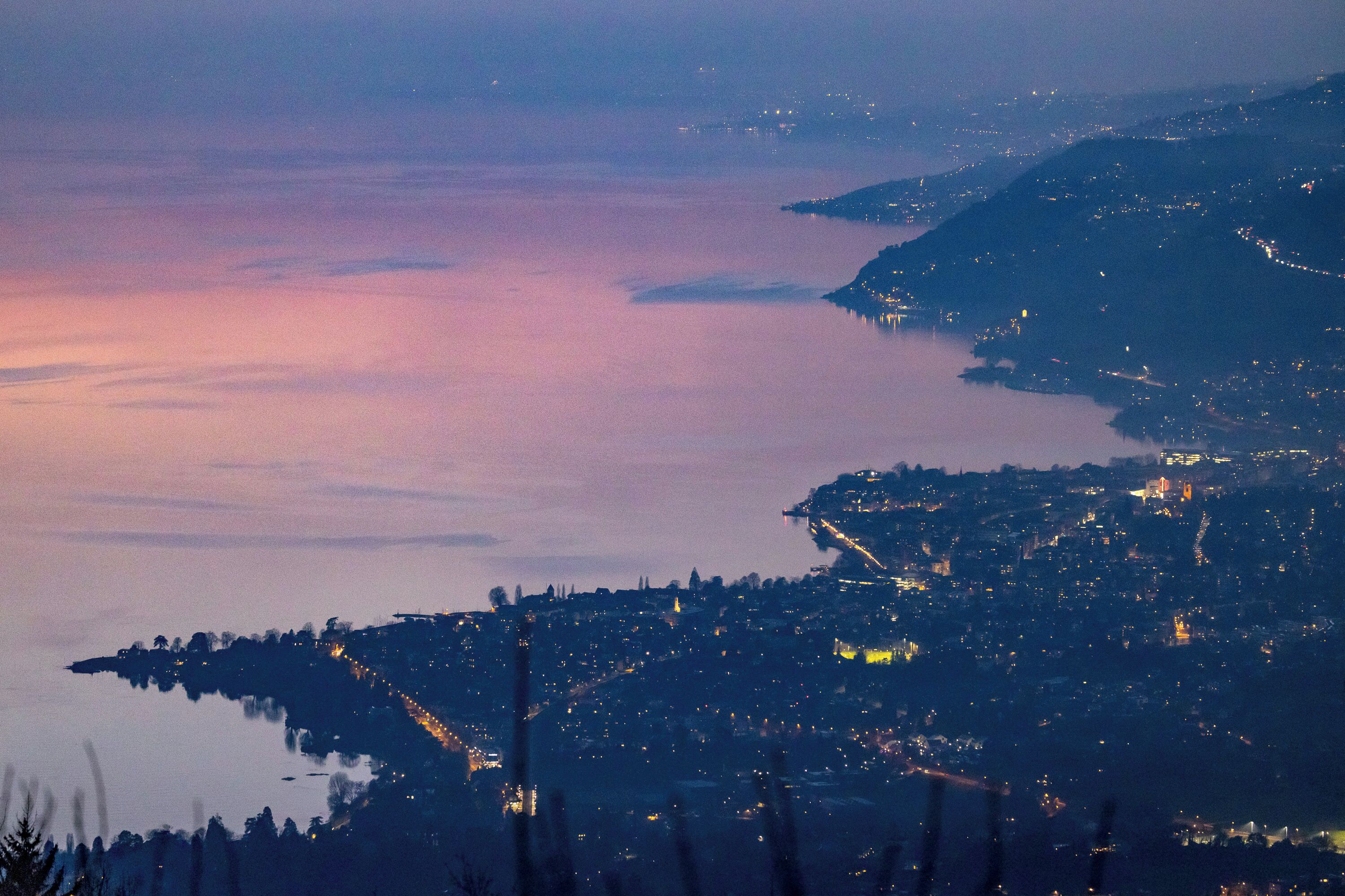 A coastal cityscape at dusk with illuminated buildings and a calm sea.