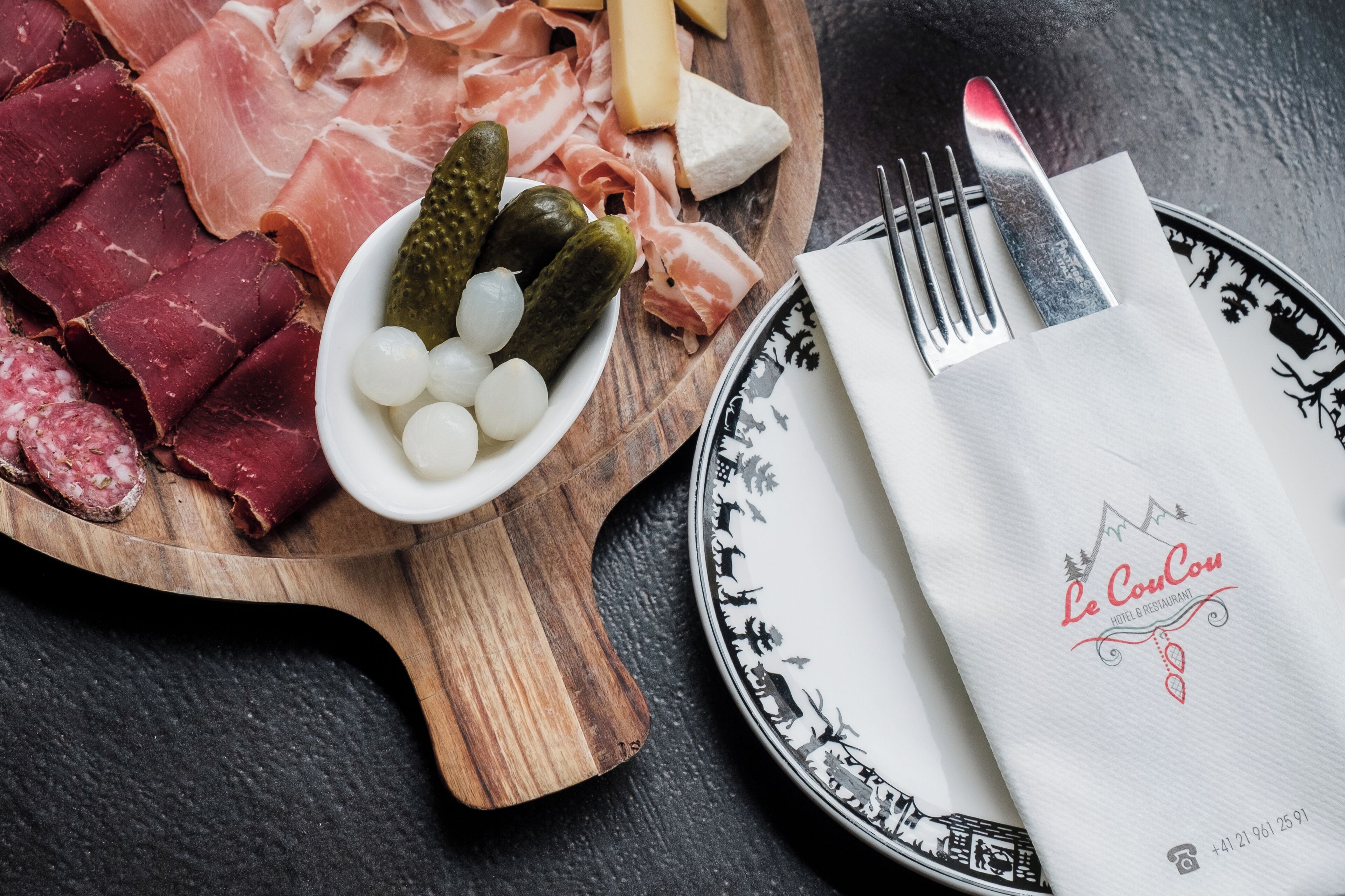 Une planche en bois garnie de différentes sortes de charcuterie, de cornichons et de fromage, une assiette avec une serviette, ainsi qu’une fourchette et un couteau.