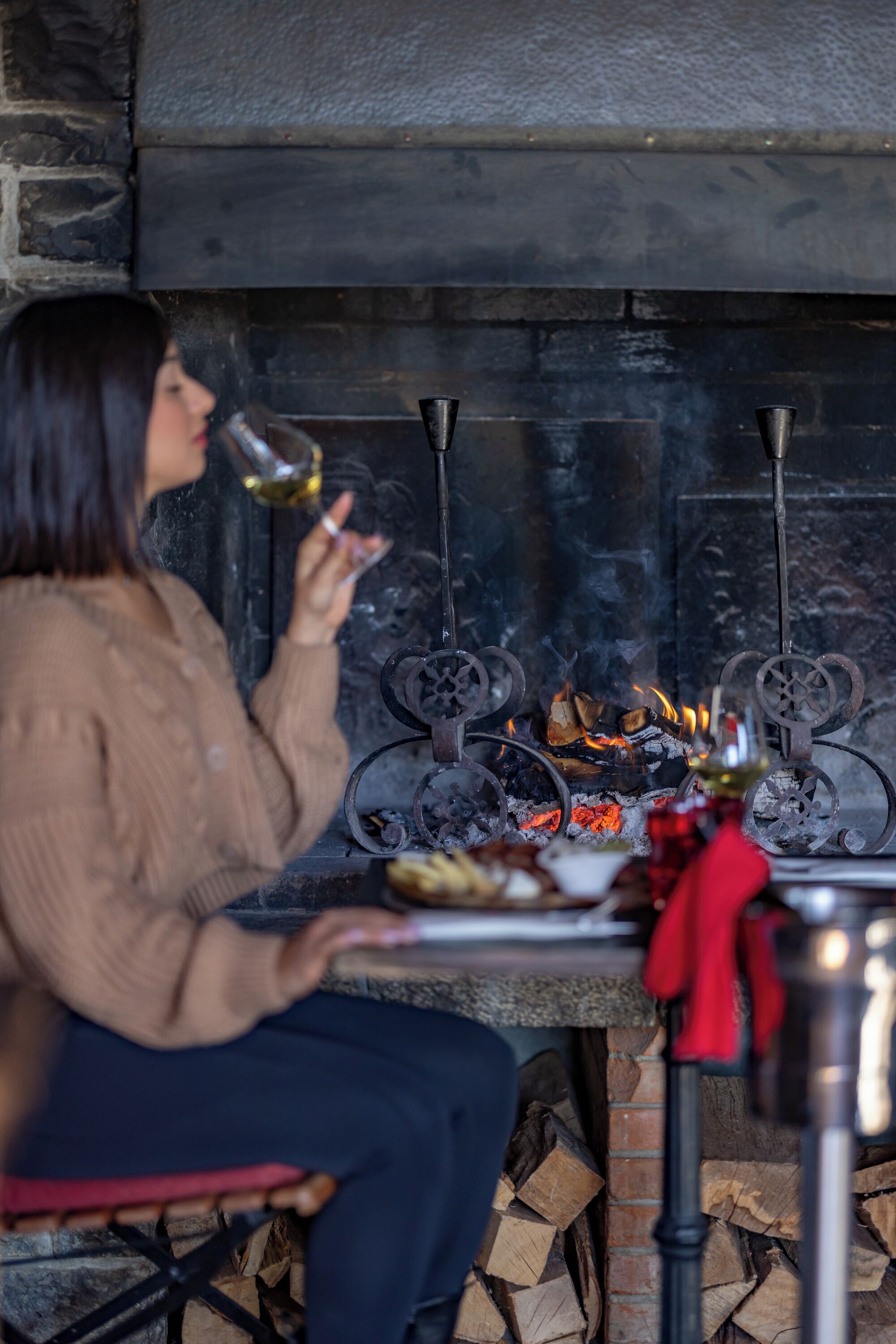 Una persona sentada a una mesa con un plato de comida y una copa de vino frente a una chimenea.