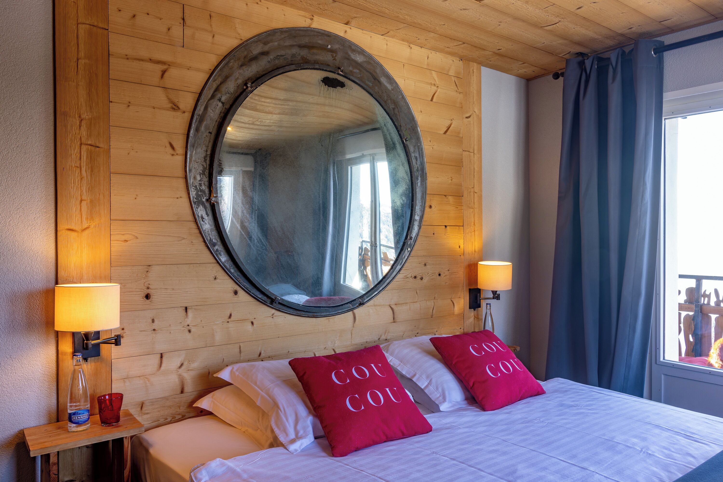 A wooden-paneled room with a round mirror, two bedside lamps, a bed with red pillows, and a window with curtains.