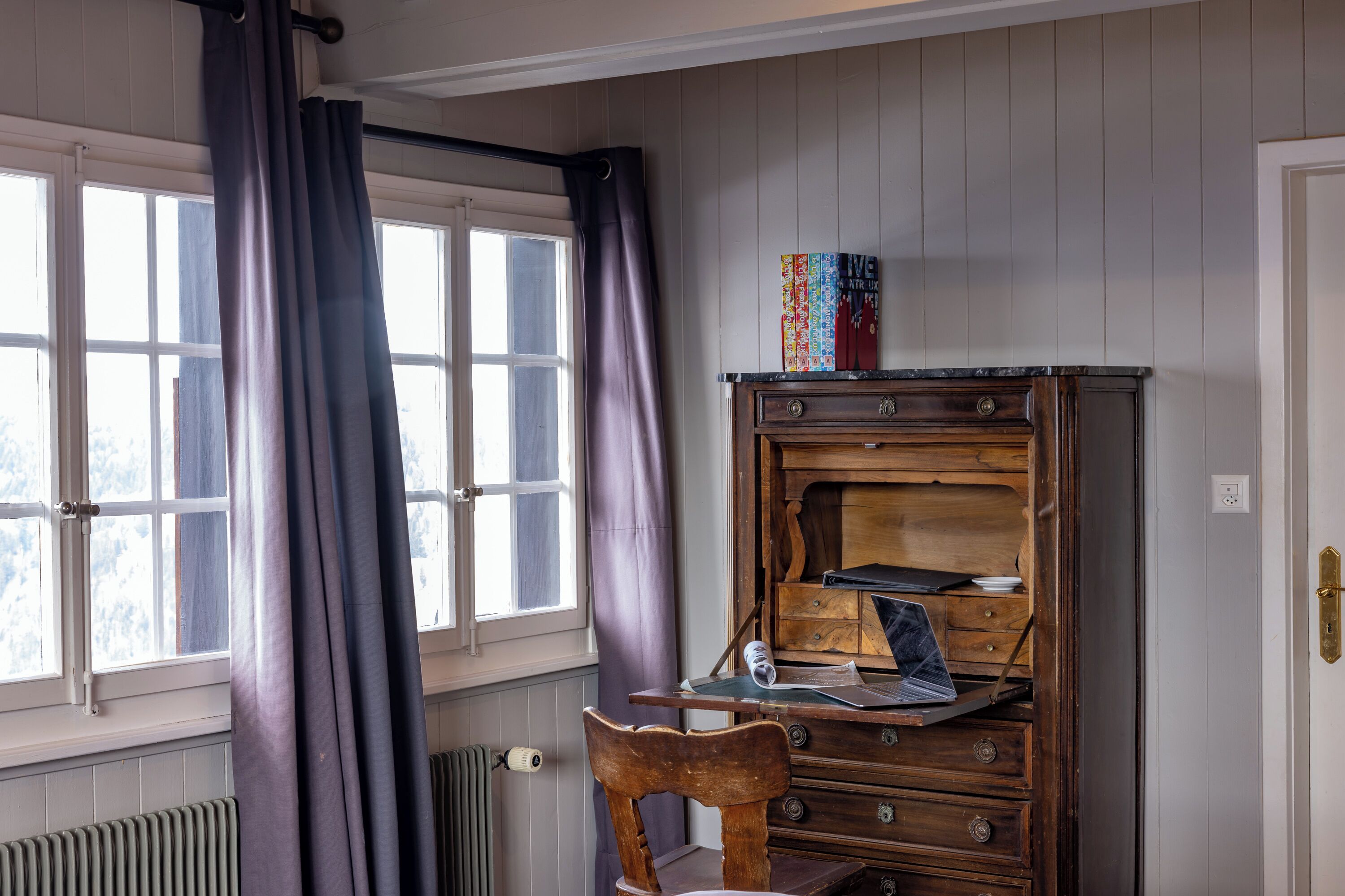 A room with a wooden dresser, a chair, a window with curtains, and a radiator.