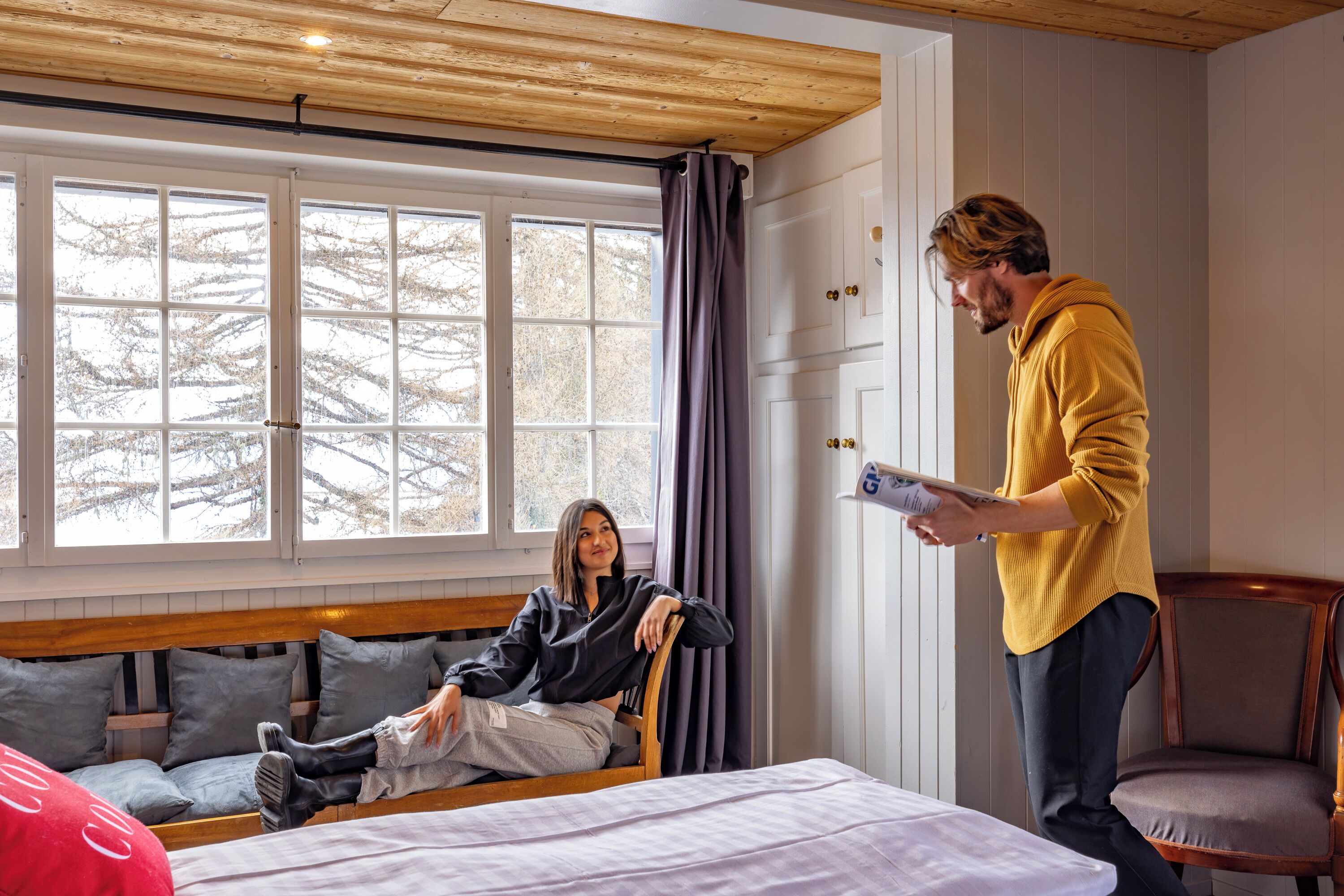 Ein Zimmer mit einem großen Fenster, einem Bett, einer Bank und einer Person, die am Fenster steht.