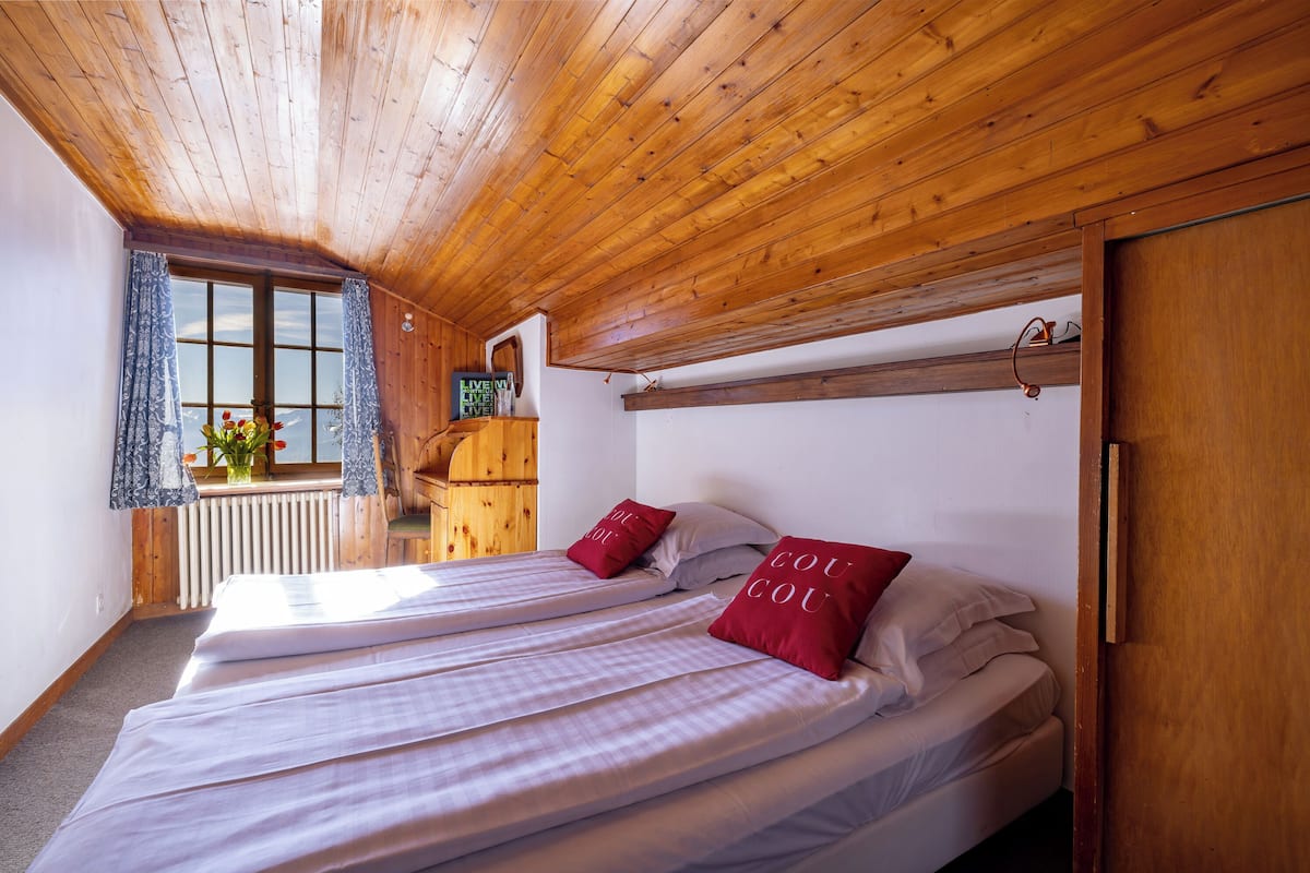 Een kamer met houten balken in het plafond, twee bedden met witte lakens en rode kussens, een raam met gordijnen en een houten kast.