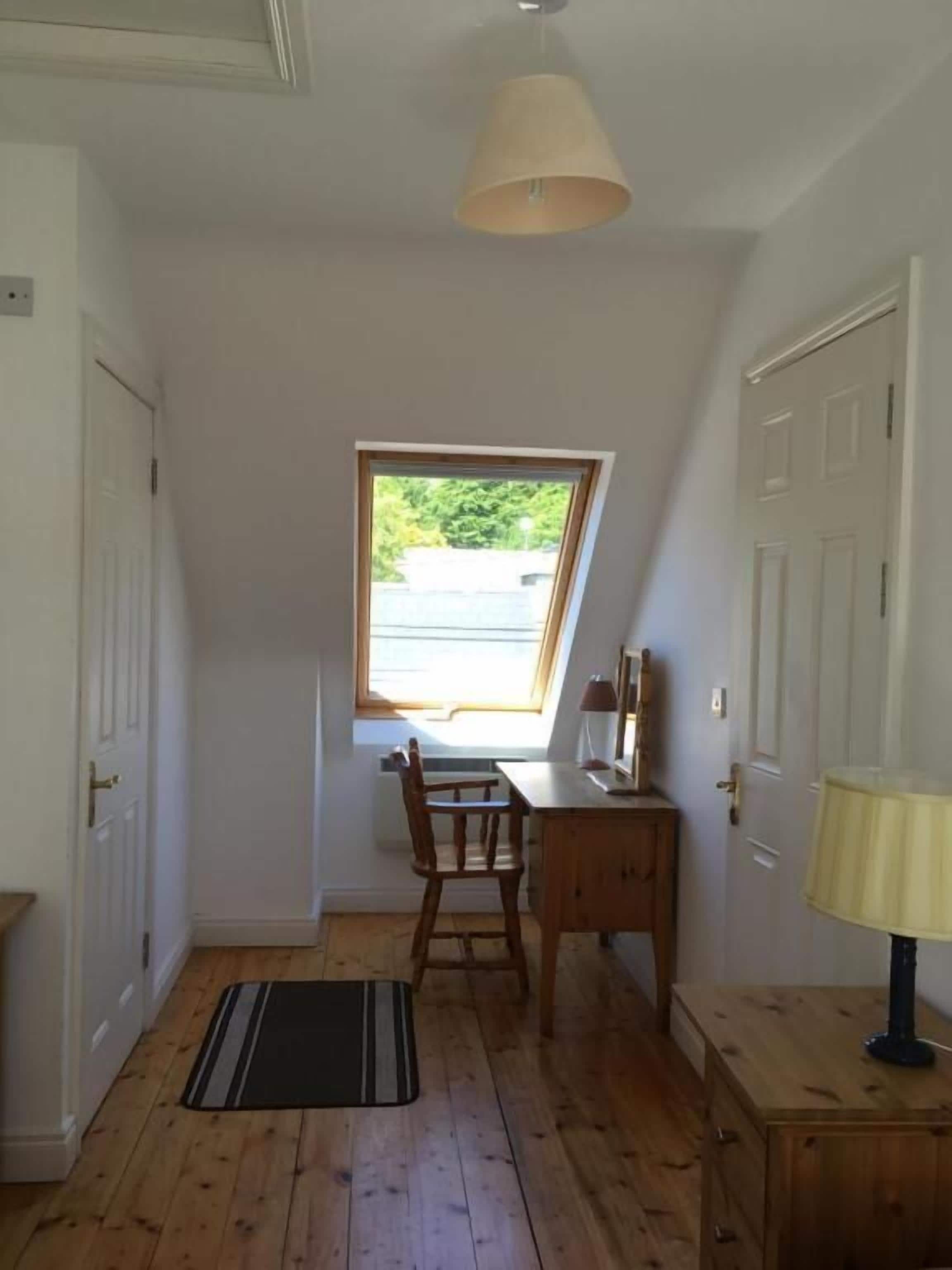 A room with a sloped ceiling, a window, a desk, a chair, a lamp, and a door.