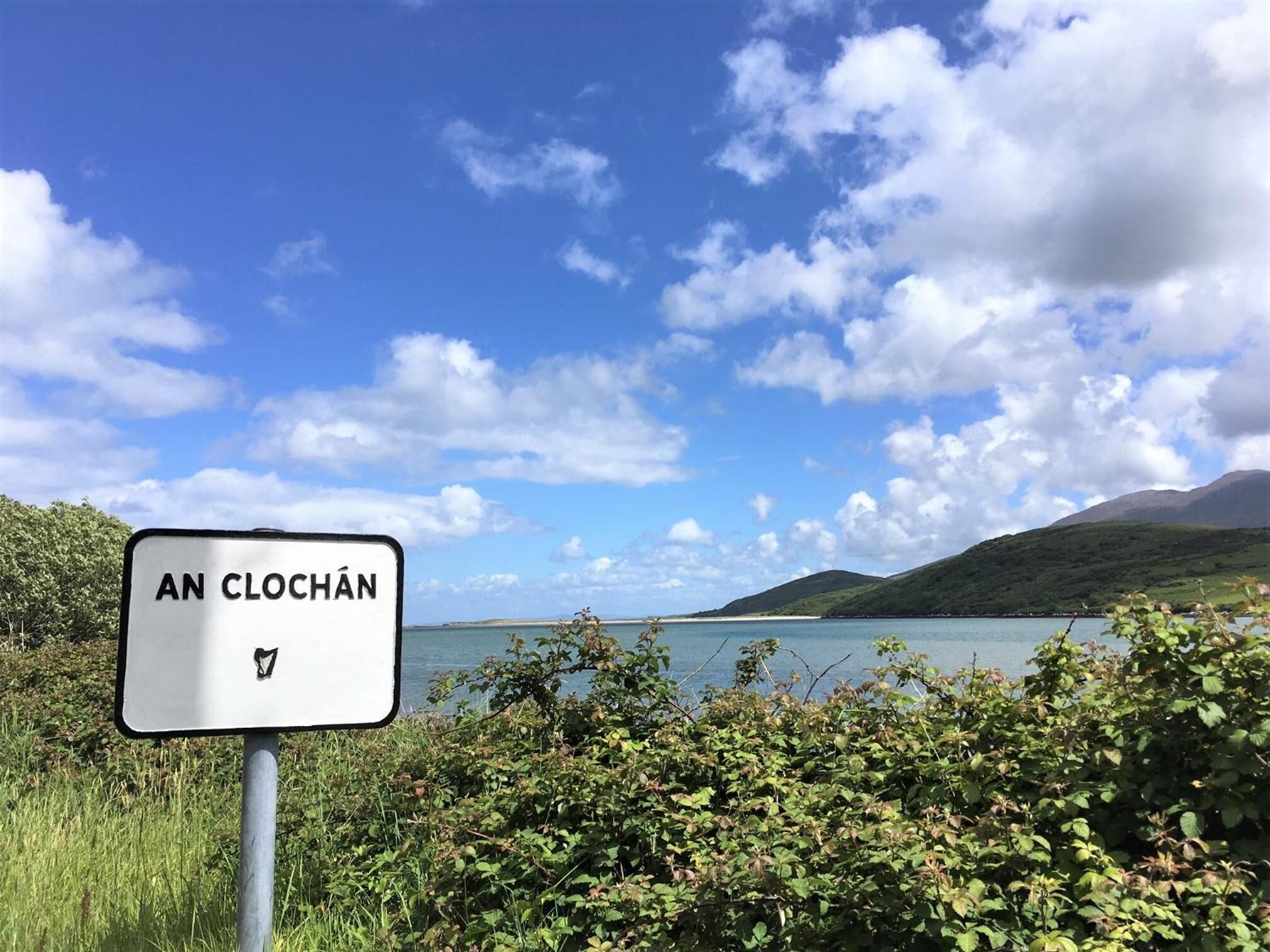 A sign with "AN CLOCHAN" written on it, surrounded by greenery and a scenic view of a lake and hills.