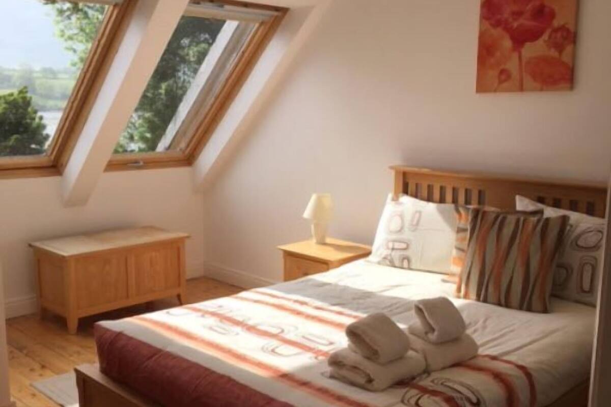 A bedroom with a wooden bed, a nightstand, a bedside table, and a window with a view of greenery.