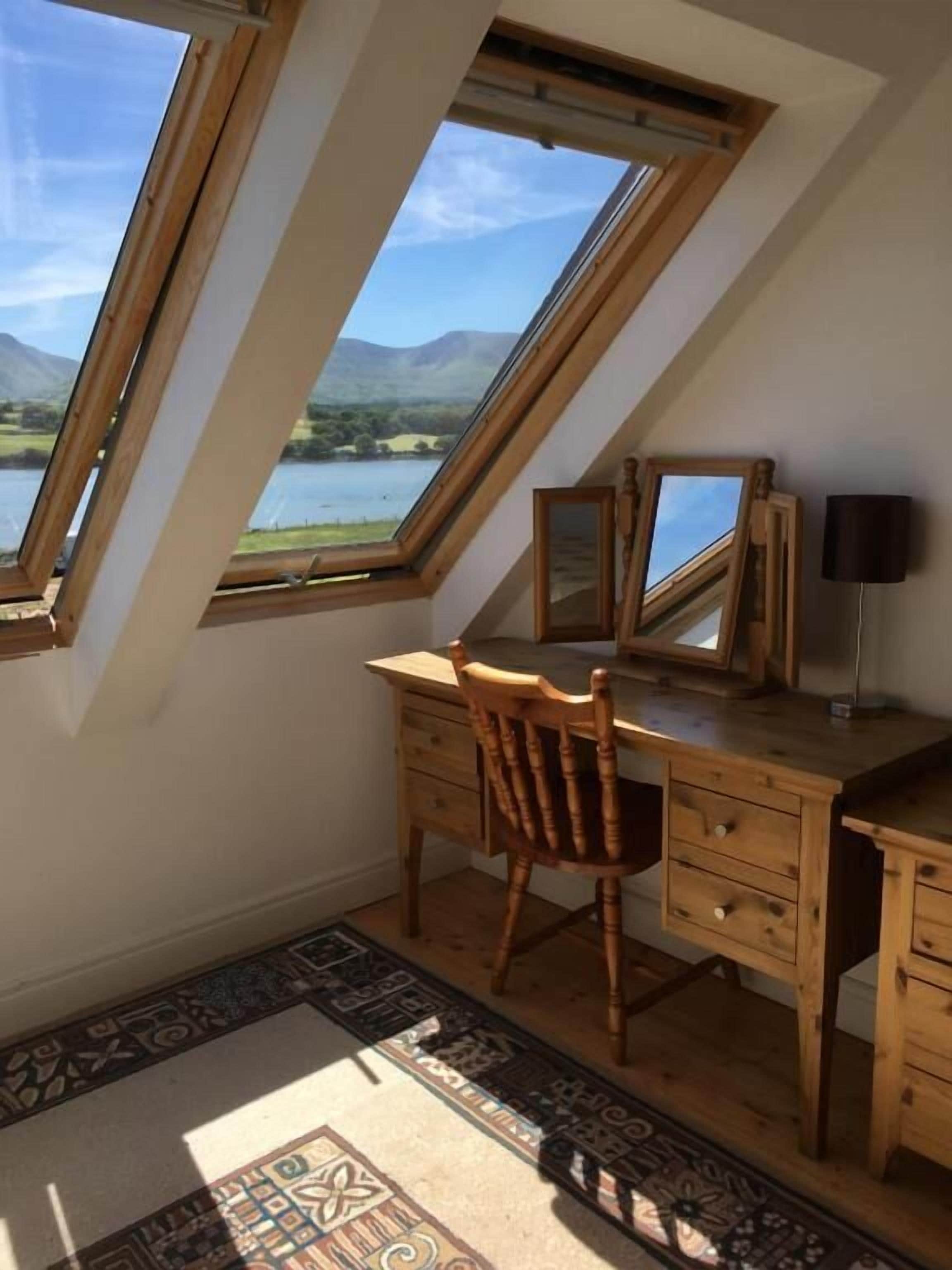 A room with a sloped ceiling, wooden desk, chair, and a view of a lake and mountains through skylights.