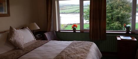 Habitación de hotel con cama, mesita de noche, silla, ventana y vistas a un paisaje verde, y un florero con flores en el alféizar.