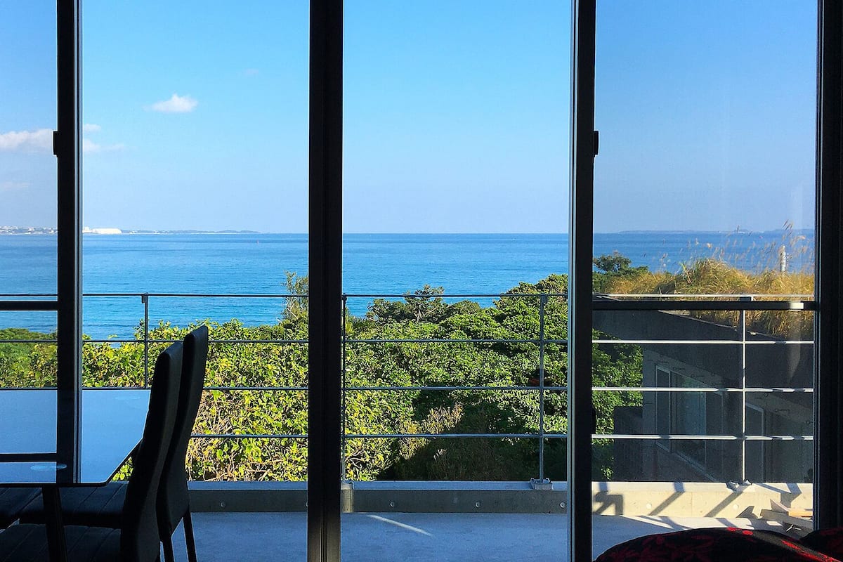 A room with a large window overlooking the sea, a balcony with a chair, and a view of distant land.