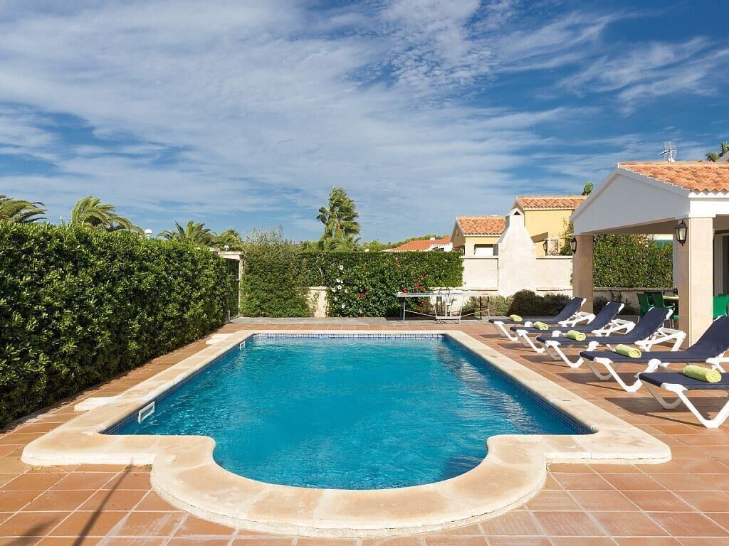 Ein rechteckiger Pool mit Fliesen umrandet, umgeben von einem niedrigen Heckenstreifen und einer Reihe von Liegestühlen mit Kissen.