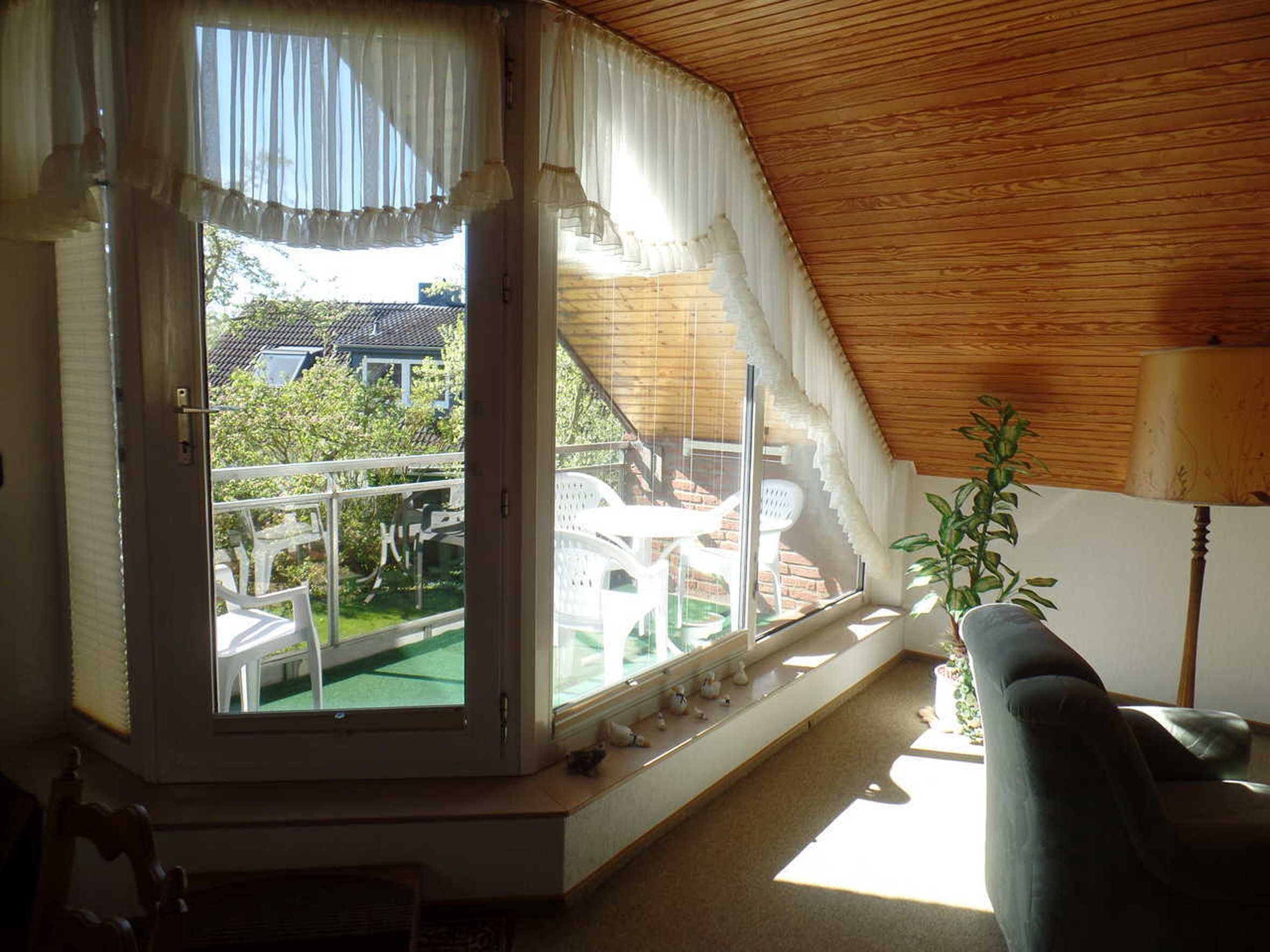 Ein Zimmer mit schrägen Wänden, einem großen Fenster mit Vorhängen, einem kleinen Tisch, einem Sessel und einer Topfpflanze.