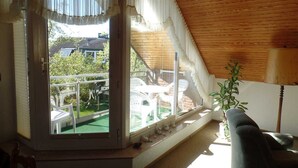 A room with a sloped ceiling, a large window with curtains, a small table, a chair, and a potted plant. - 3 room apartment 80sqm - Apartments Eggert (Neustadt in Holstein)