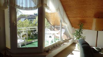 Ein Zimmer mit schrägen Wänden, einem großen Fenster mit Vorhängen, einem kleinen Tisch, einem Sessel und einer Topfpflanze.