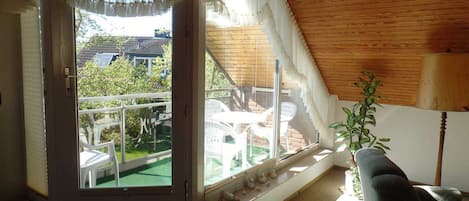Una habitación con techo inclinado, un ventanal con cortinas, una mesa pequeña, una silla y una planta en maceta.