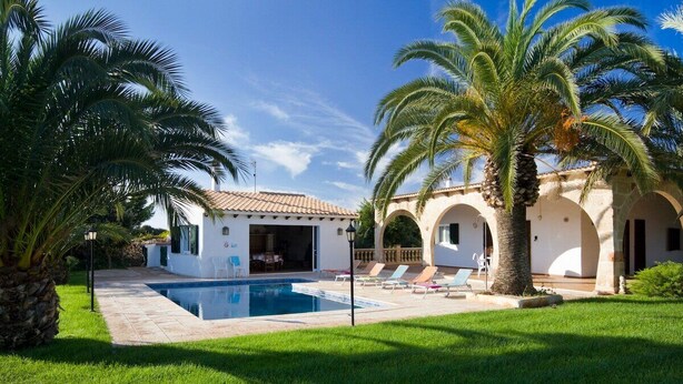 A poolside area with a clear blue pool, palm trees, and a white building with arches.