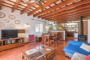 A cozy living room with a TV, wooden furniture, a dining area, and a kitchen with a brick wall and a wood stove.