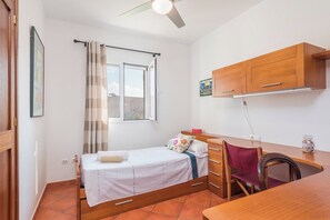 Una stanza piccola e luminosa con un letto, una scrivania e una sedia. Si vede una finestra con tende, una ventola a soffitto e un mobiletto a parete.