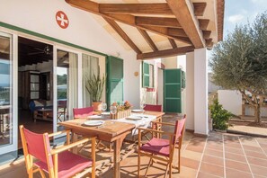 Un'area pranzo all'aperto con tavoli e sedie di legno, persiane verdi e vista sull'interno.
