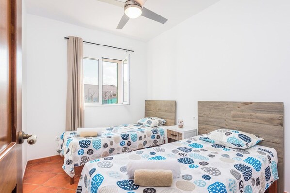 Une chambre d’hôtel avec deux lits, une tête de lit en bois, une fenêtre avec des rideaux et un ventilateur de plafond.