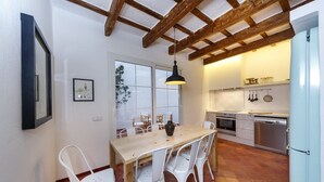 A dining area with a wooden table and white chairs, a hanging light fixture, and a kitchen with white cabinets and stainless steel appliances. - Sant Isidre 50, 4 bedroom house, Ciutadella (Ciutadella de Menorca)