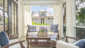 A covered porch with a wooden bench and chairs, a table with a basket of flowers, and a view of a house with a white fence and greenery. - Updated Wilson Village 4 Bedroom Cottage  Amenity access available (Bluffton)