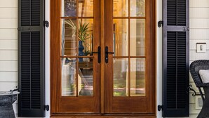 A wooden double door with glass panels, flanked by black shutters, leading to a porch with a doormat and a chair. - Updated Wilson Village 4 Bedroom Cottage  Amenity access available (Bluffton)