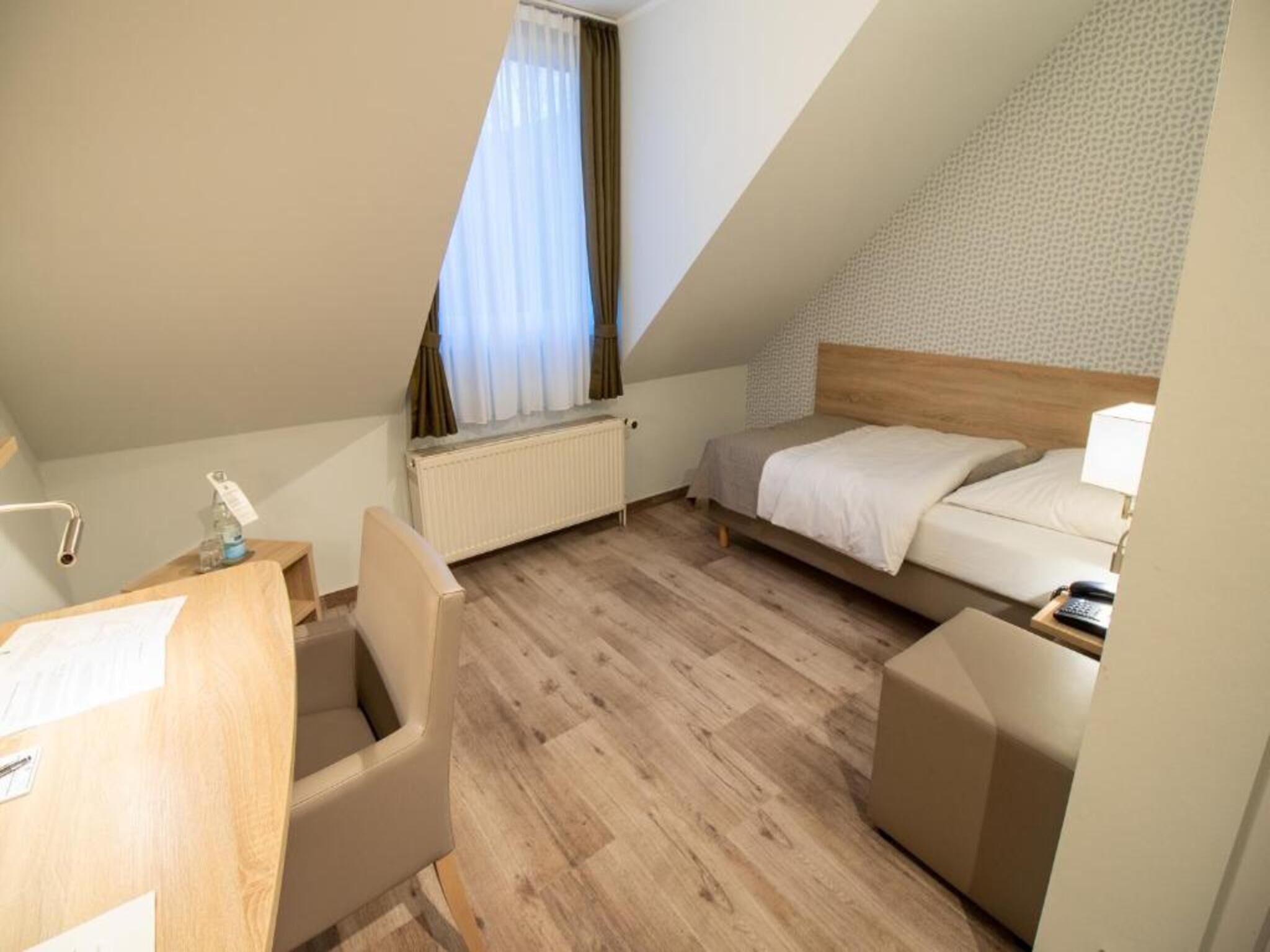 A small, single-room hotel room with a wooden floor, a bed, a desk with a chair, a radiator, and a window with curtains.