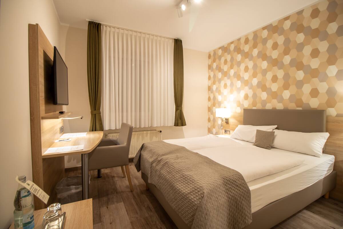 Ett hotellrum med en säng, ett skrivbord, en stol, en tv och ett fönster med gardiner.