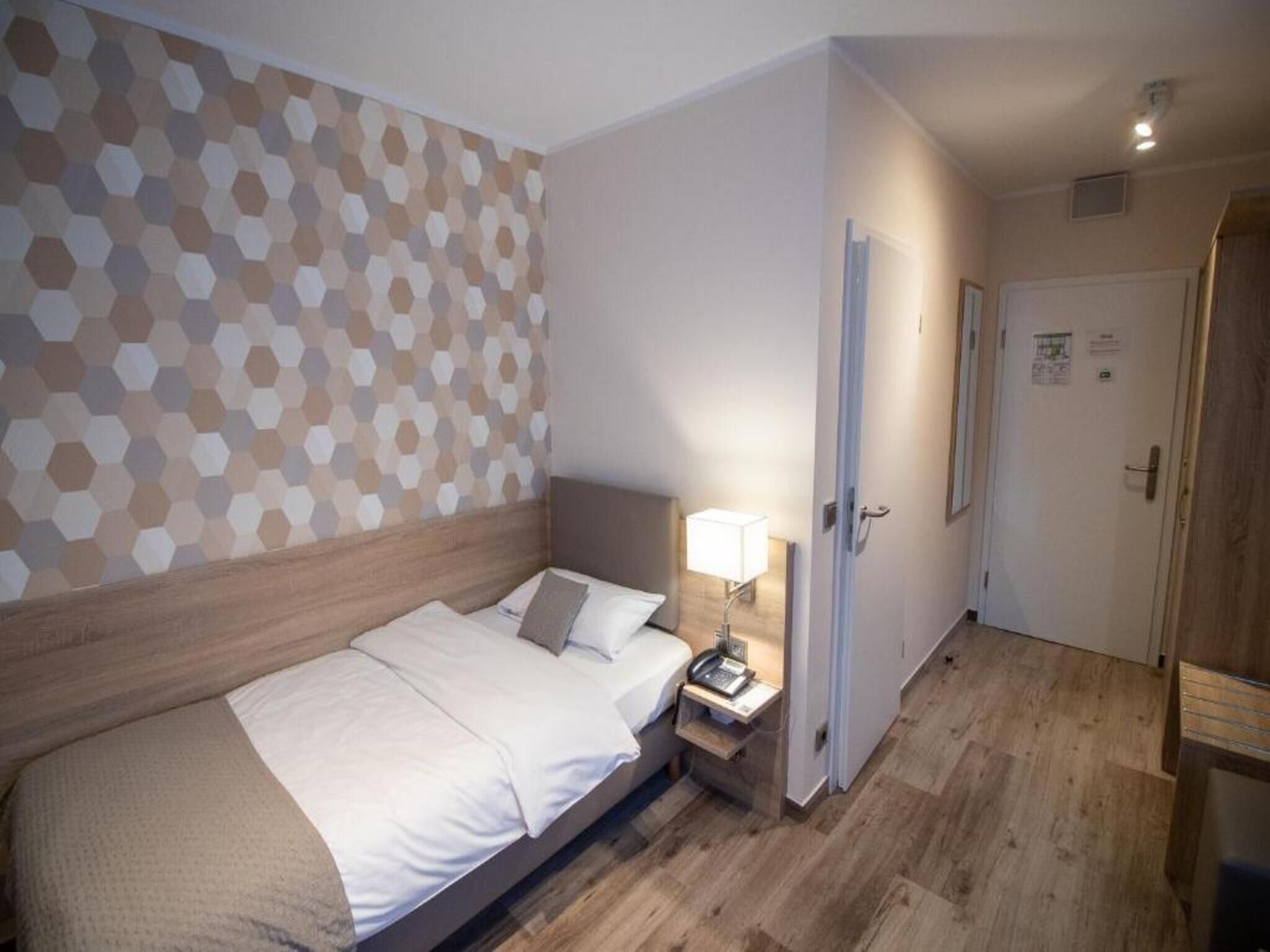 A hotel room with a bed, bedside table with a lamp, a door, and a wall with a geometric pattern.