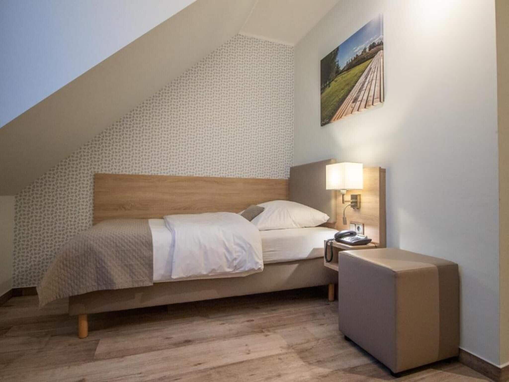 Un dormitorio con techo inclinado, una cama con cabecera de madera, una mesita de noche con lámpara y un cuadro en la pared.