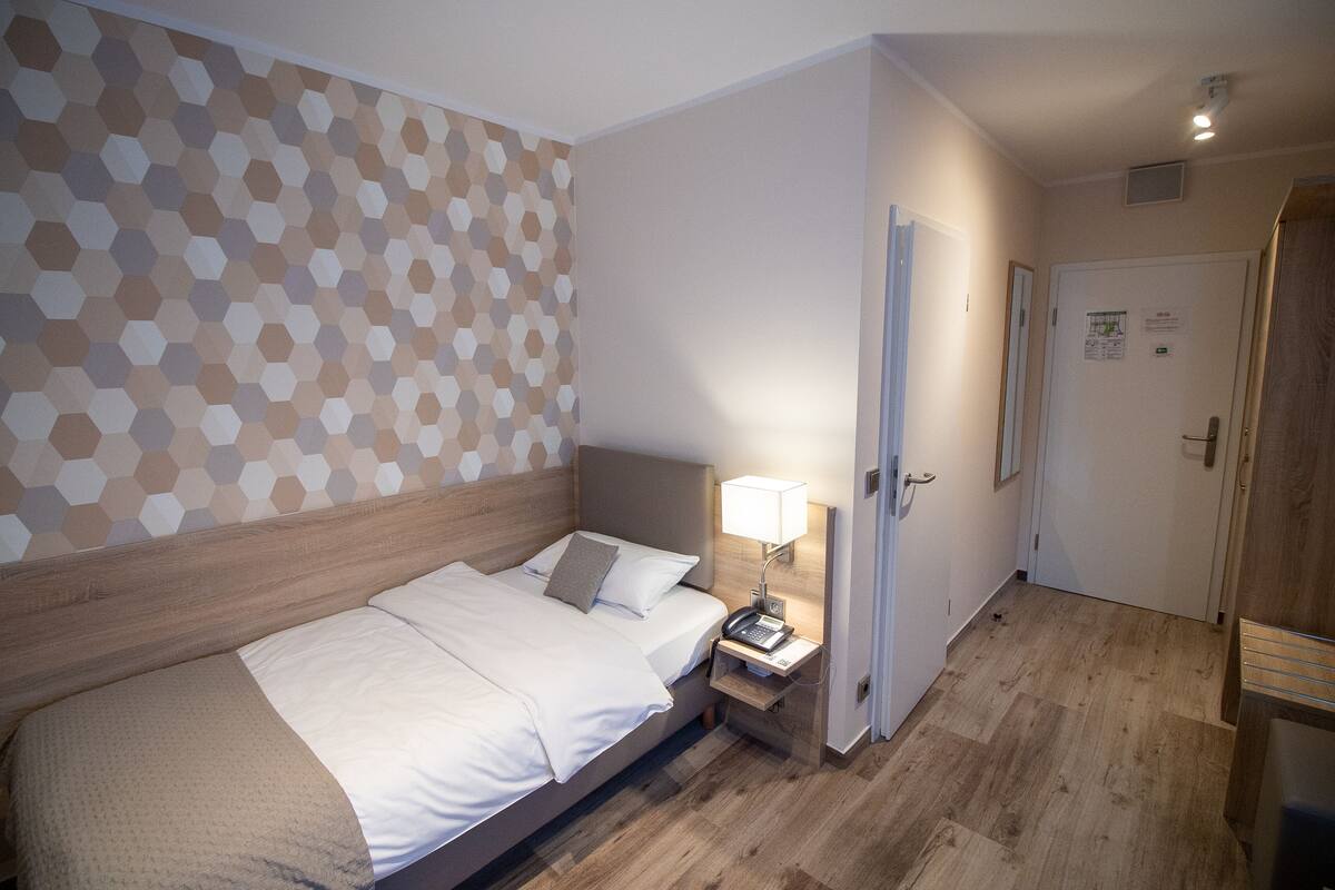 Ett hotellrum med en sänggavel i trä, en säng med vita sängkläder, ett nattduksbord med en lampa och en vägg med ett geometriskt mönster.
