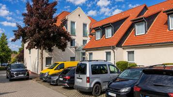 A row of parked cars in front of a building with a red-tiled roof and a tree with red leaves.