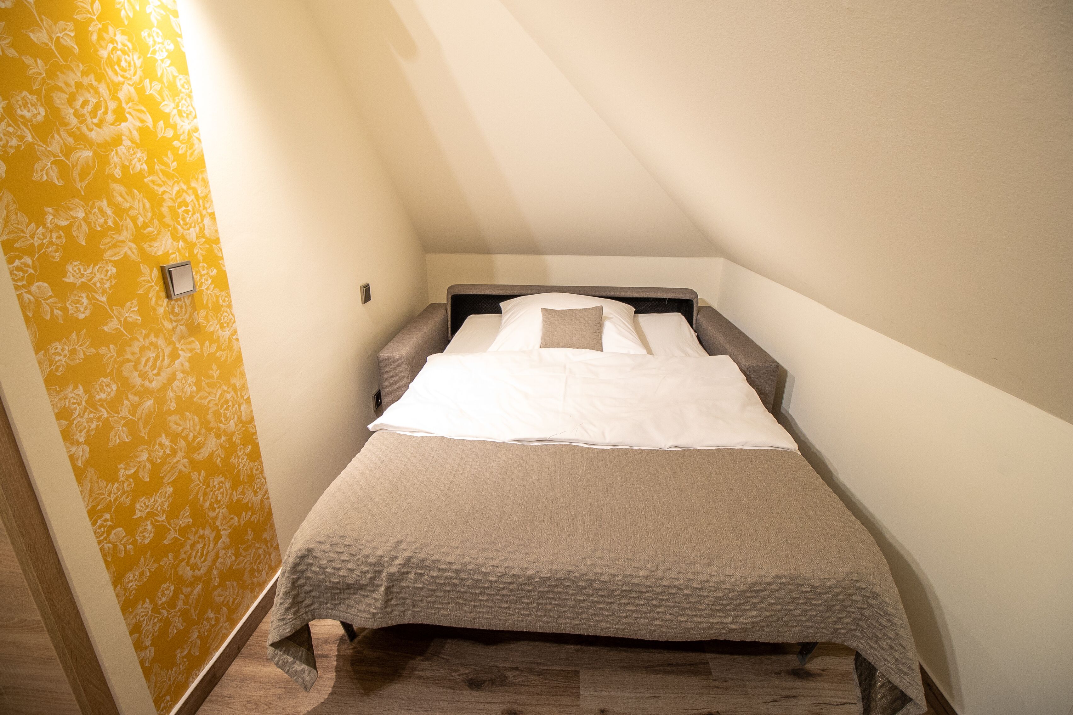Una cama pequeña y ordenada en una habitación de esquina, con piso de madera y un papel tapiz floral amarillo en una pared.