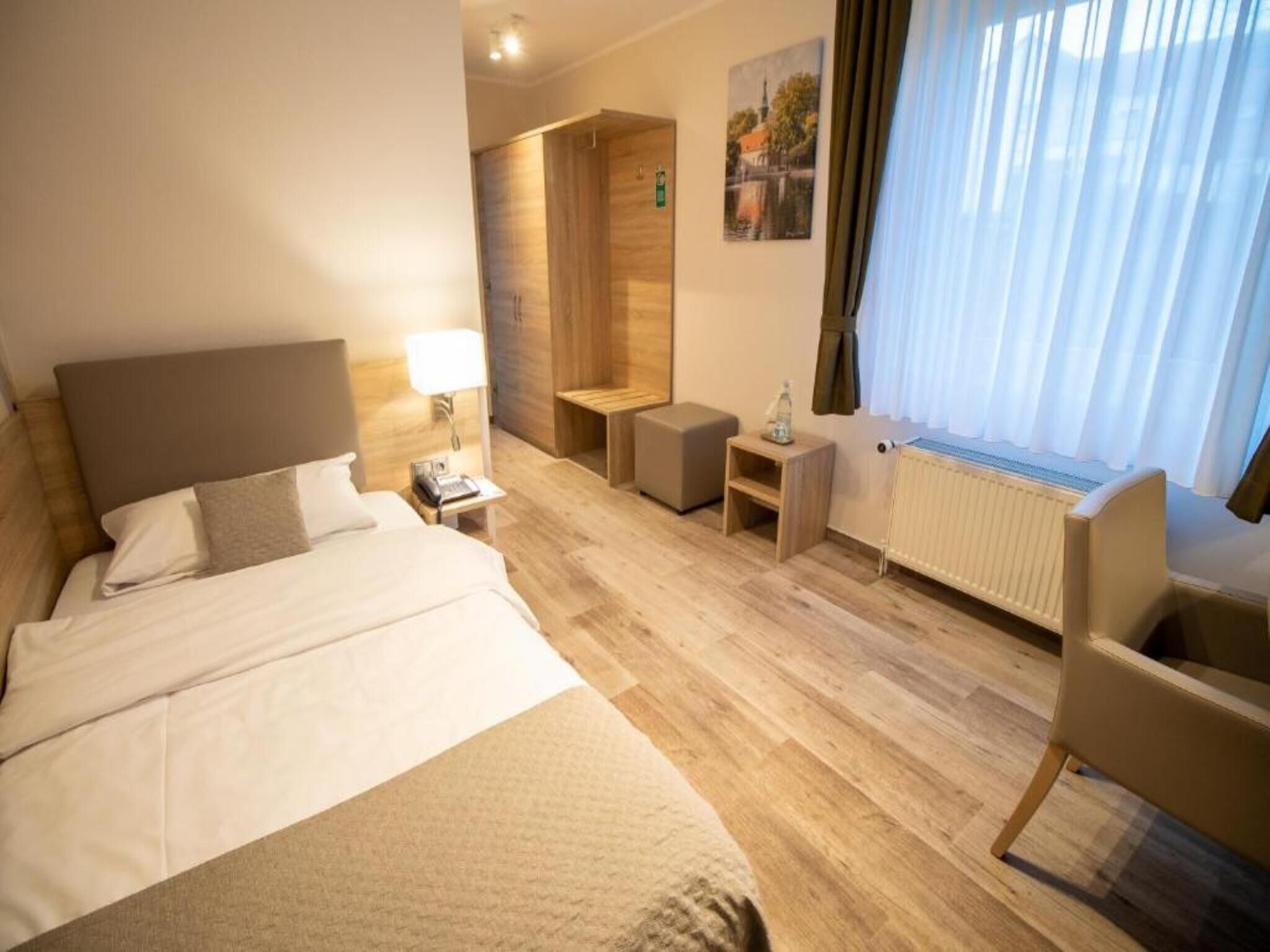 Ein Hotelzimmer mit Bett, Nachttisch, Lampe, einer kleinen Holzbank, einem Sessel und einem Heizkörper.