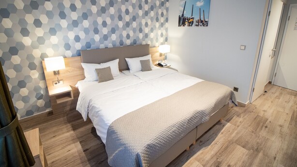 Et moderne hotelværelse med en stor seng, to sengeborde med lamper, en væg med geometrisk mønster og et bybillede som maleri.