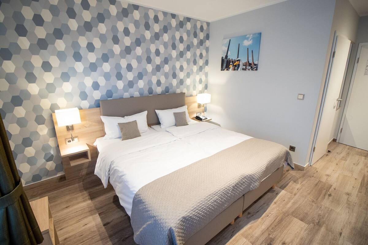 Ett modernt hotellrum med en stor säng, två sängbord med lampor, en vägg med ett geometriskt mönster och en tavla som föreställer en stadsbild.