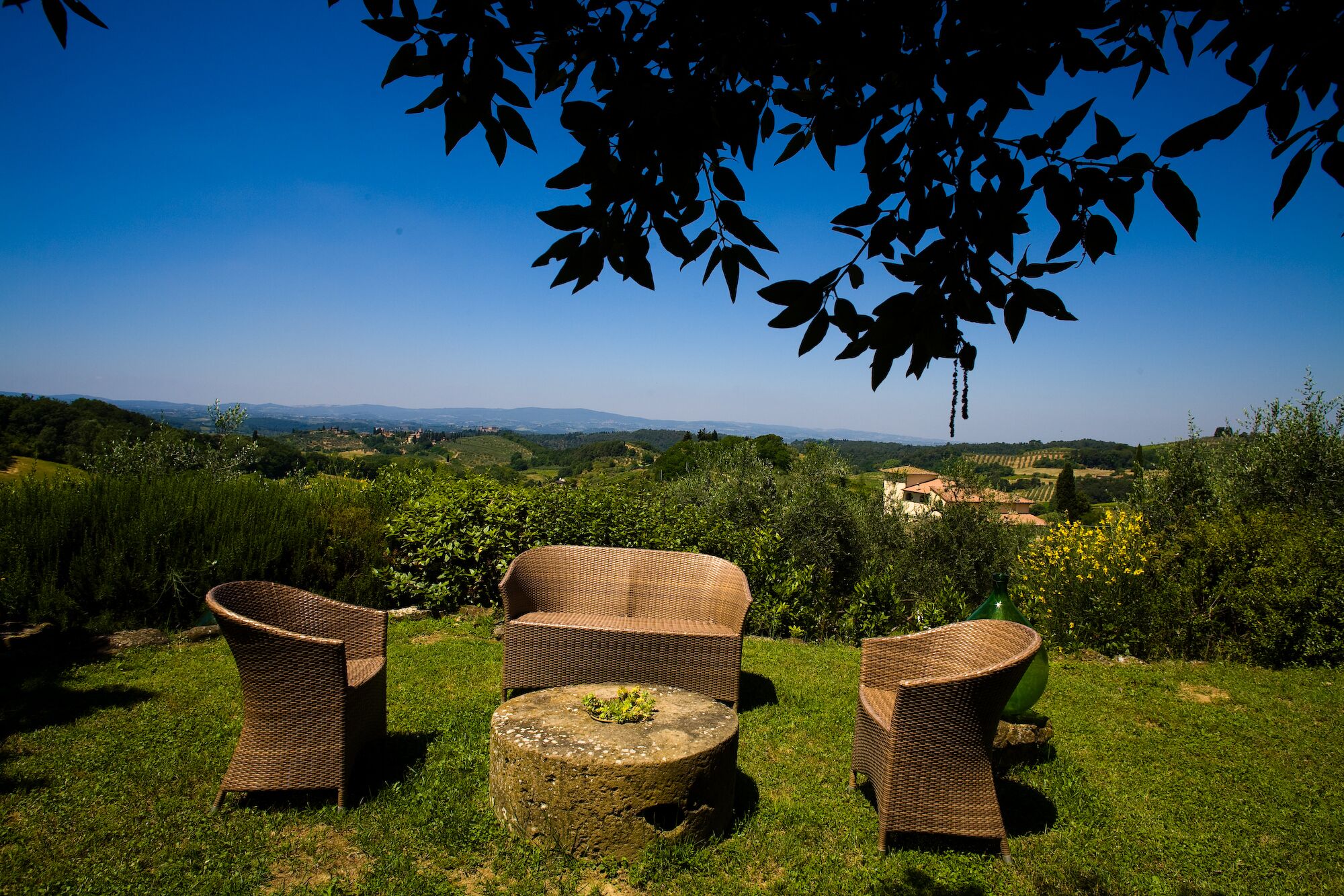 A wicker outdoor seating area with a stone table, surrounded by lush greenery and a scenic view of distant hills and buildings.