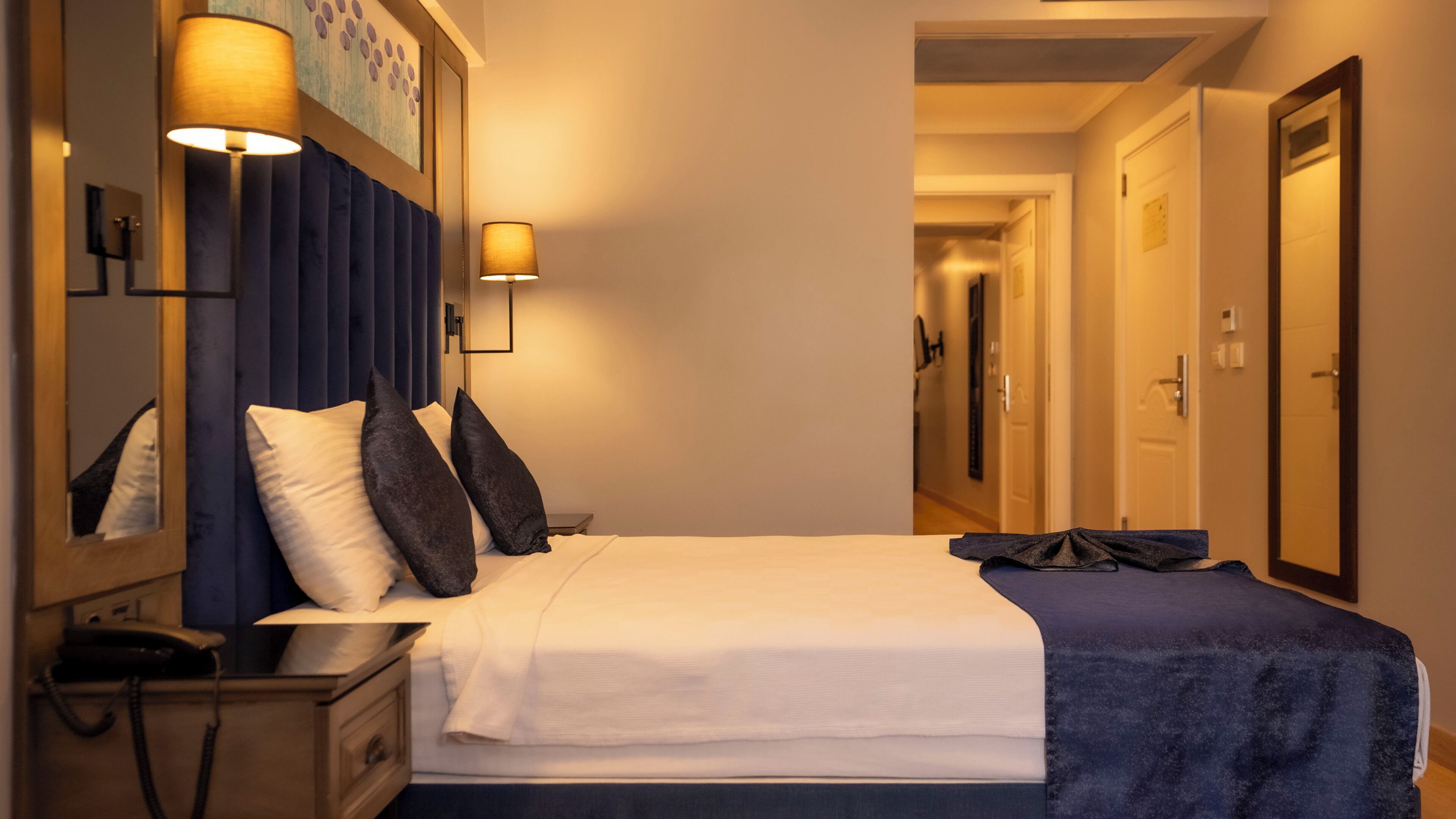 Una habitación de hotel con cama, lámparas de noche, mesita de noche, espejo y puerta.