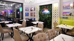 Un restaurante con mesas de mármol y sillas acolchadas, una araña y una pared decorada con cuadros enmarcados y un fondo de plantas verdes.