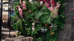 Un vestíbulo con una pared verde decorada con plantas y flores, un sofá marrón con textura y una mesa de mármol.