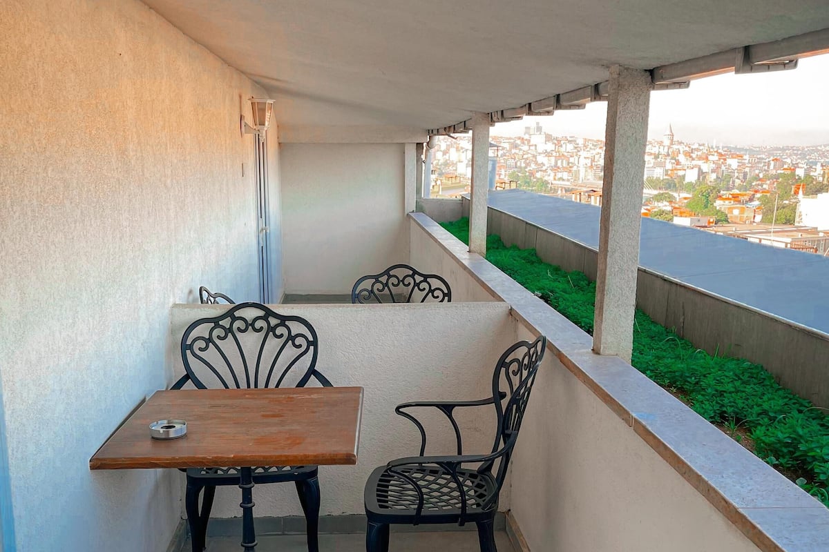 A balcony with a table and two chairs, a view of a cityscape, and a green area along the railing.