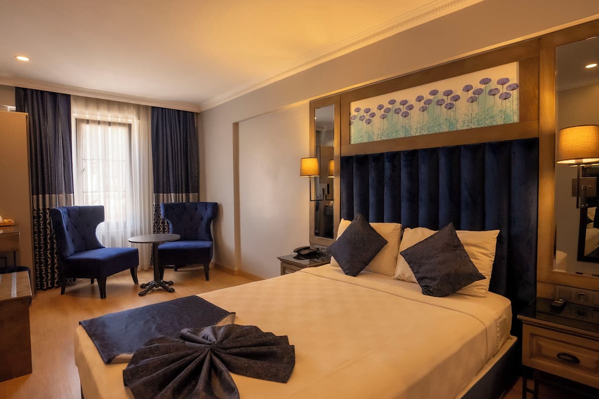 A hotel room with a large bed, two armchairs, a small table, a nightstand with a lamp, and a painting on the wall.