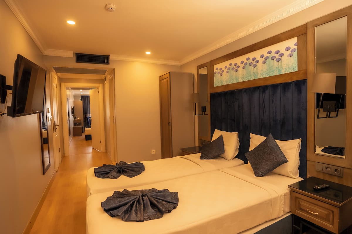 A hotel room with a large bed, a flat-screen TV mounted on the wall, a bedside table with a lamp, and a large mirror above the bed.