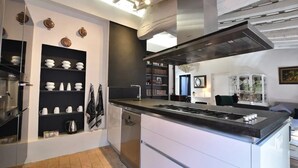Uma cozinha moderna com armários brancos, bancada preta e fogão embutido. Há prateleiras com louças brancas e uma máquina de café.