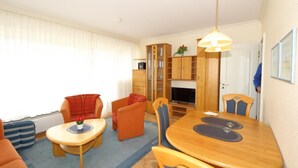 A compact living space with a dining table, two sofas, a TV, and a bookshelf. - Becker House Alice 36 (Sylt)