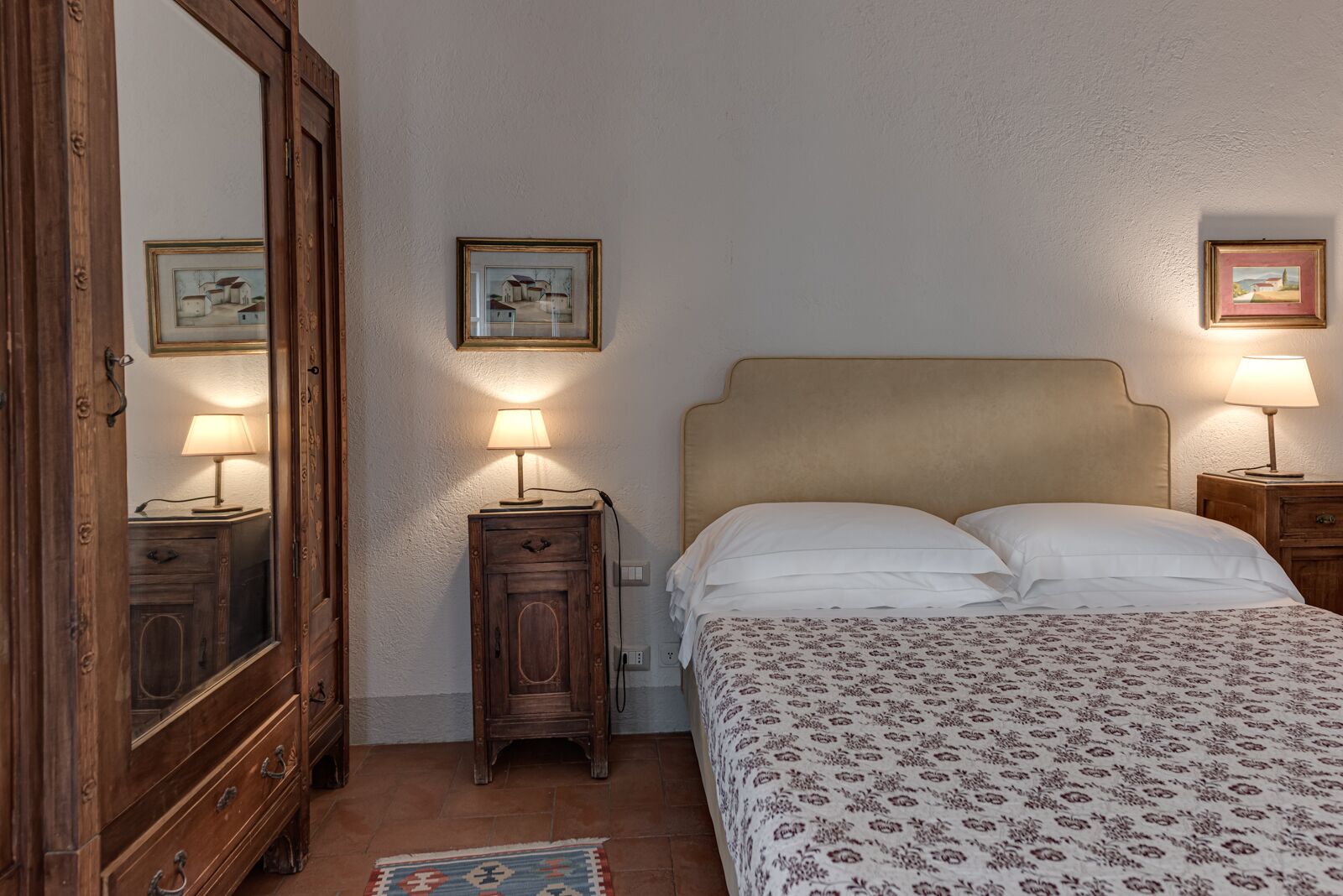 A bedroom with a bed, two bedside tables with lamps, a wooden wardrobe, and two framed pictures on the wall.