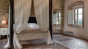 Ein Schlafzimmer mit einem Himmelbett, Steinwänden, einem großen Spiegel und Blick durch ein Fenster.