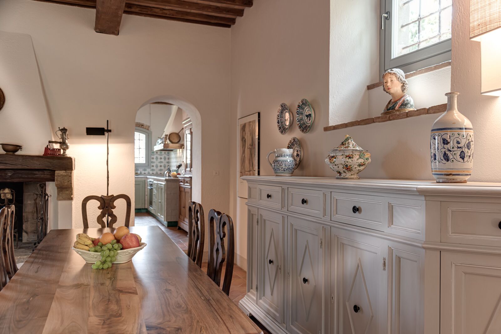 A dining area with a wooden table, chairs, and a fruit bowl. There is a white cabinet with decorative plates and a vase. A fireplace is visible in the background.